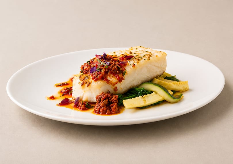 Pan-seared fish with red pepper relish, zucchini, and spinach on a white plate.