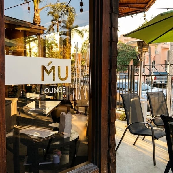 Exterior view of MÚ LOUNGE with outdoor patio seating under a green umbrella.
