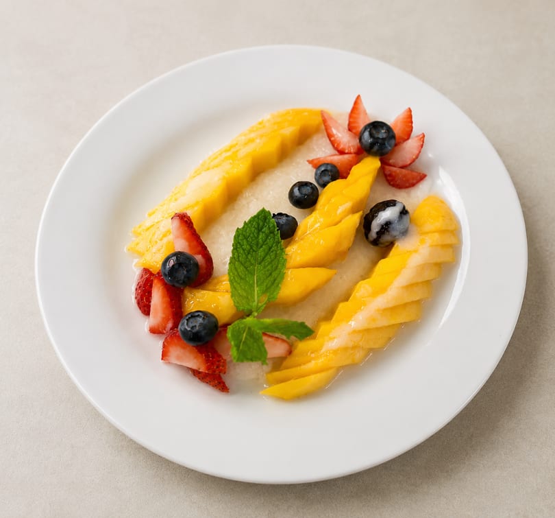 Sliced mangoes, strawberries, blueberries, and mint leaves arranged on a white plate with sauce.