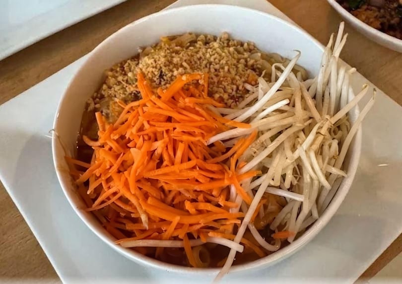 A white bowl of Pad Thai noodles topped with shredded carrots, bean sprouts, and crushed peanuts.