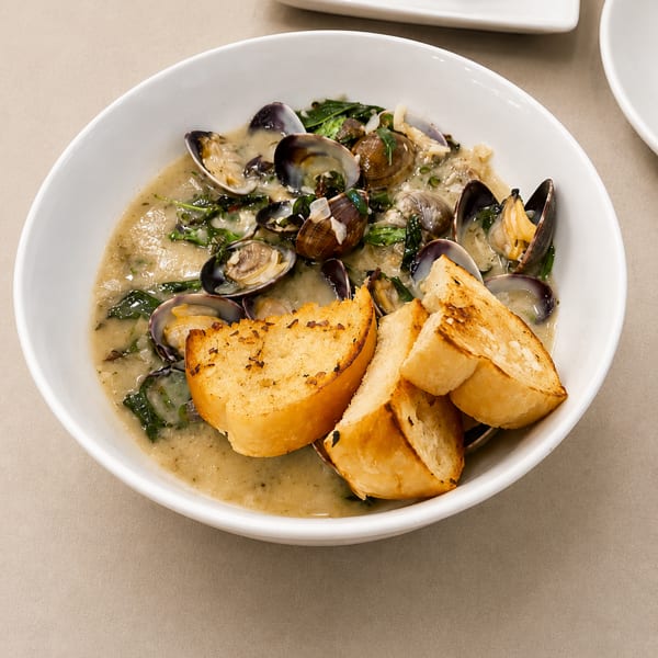 Clams in a creamy sauce with greens and toasted bread in a white bowl.